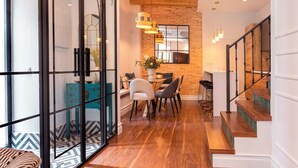Un interior moderno con una escalera de madera, un comedor con mesa y sillas, y una cocina con pared de ladrillo y lámparas colgantes.