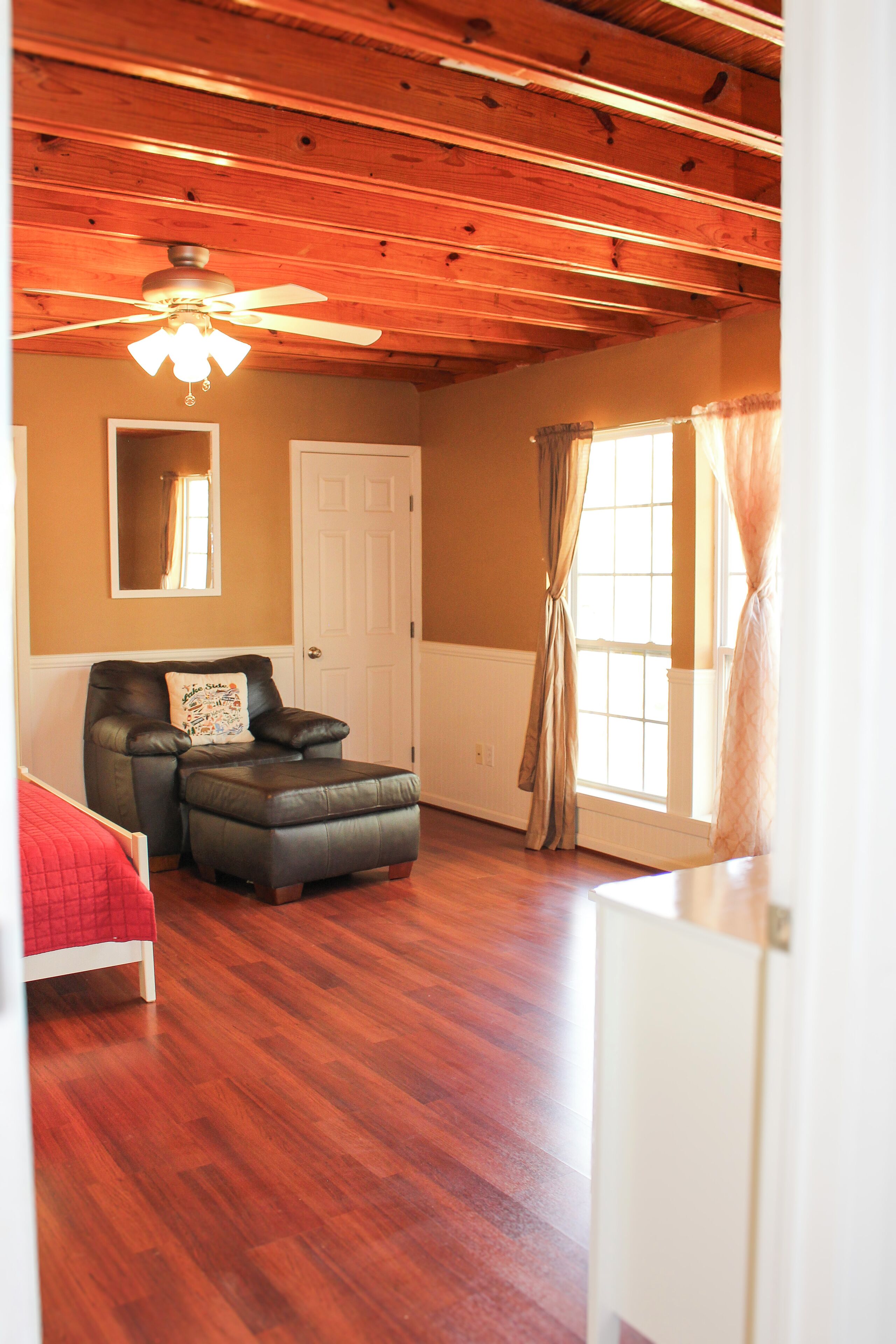 Une pièce avec un plafond en bois, un ventilateur de plafond, une fenêtre avec des rideaux, une porte, une chaise avec un coussin et un lit recouvert d’une couverture rouge.