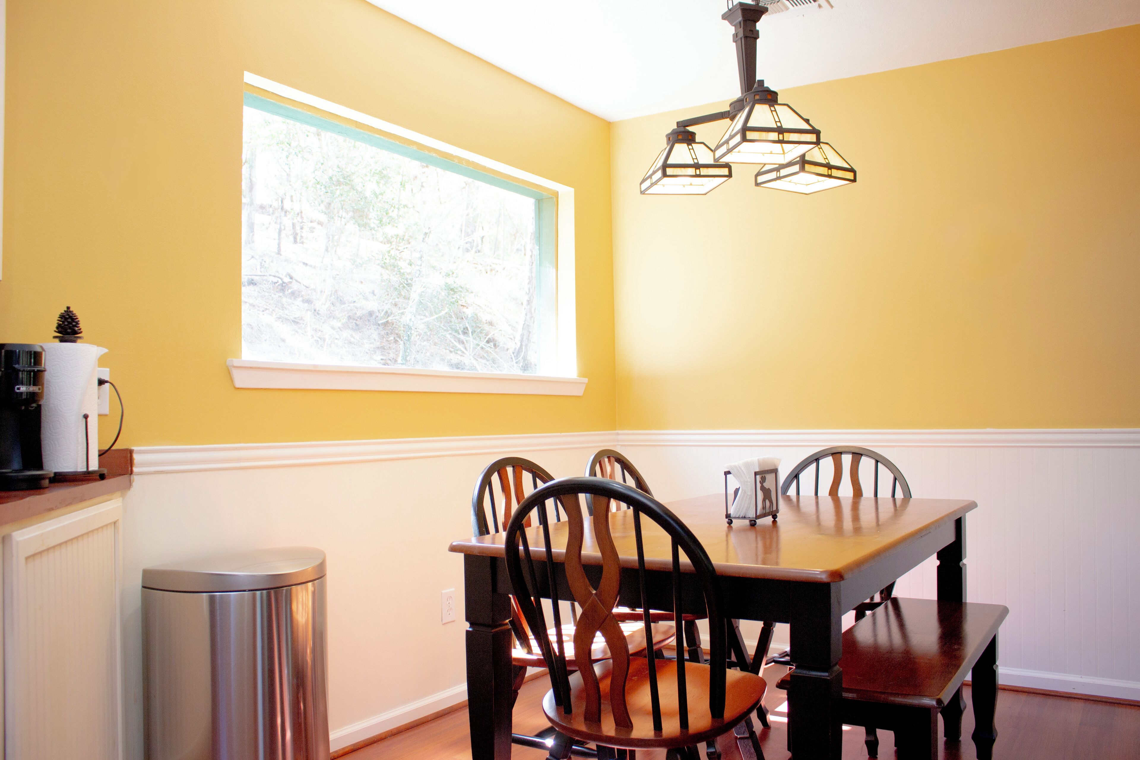 A dining area with a wooden table and chairs, a window with a view of trees, and a hanging light fixture.