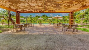 An outdoor patio area with tables and chairs, a pergola overhead, and a view of a pool and greenery. - PF77 - Apartment in Vila do Lago Cond with pool (Mata de São João)