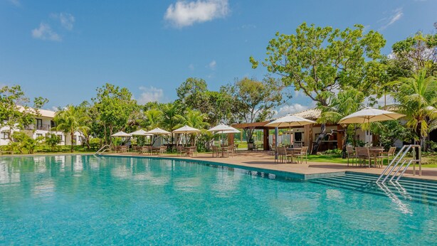 A clear blue pool with lounge chairs and umbrellas, surrounded by greenery and buildings. - PF77 - Apartment in Vila do Lago Cond with pool (Mata de São João)