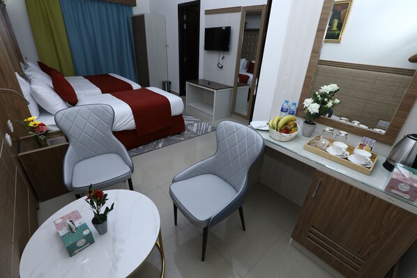 A hotel room with a bed, two chairs, a small table with a vase, a tray with cups and a banana, and a TV on the wall.