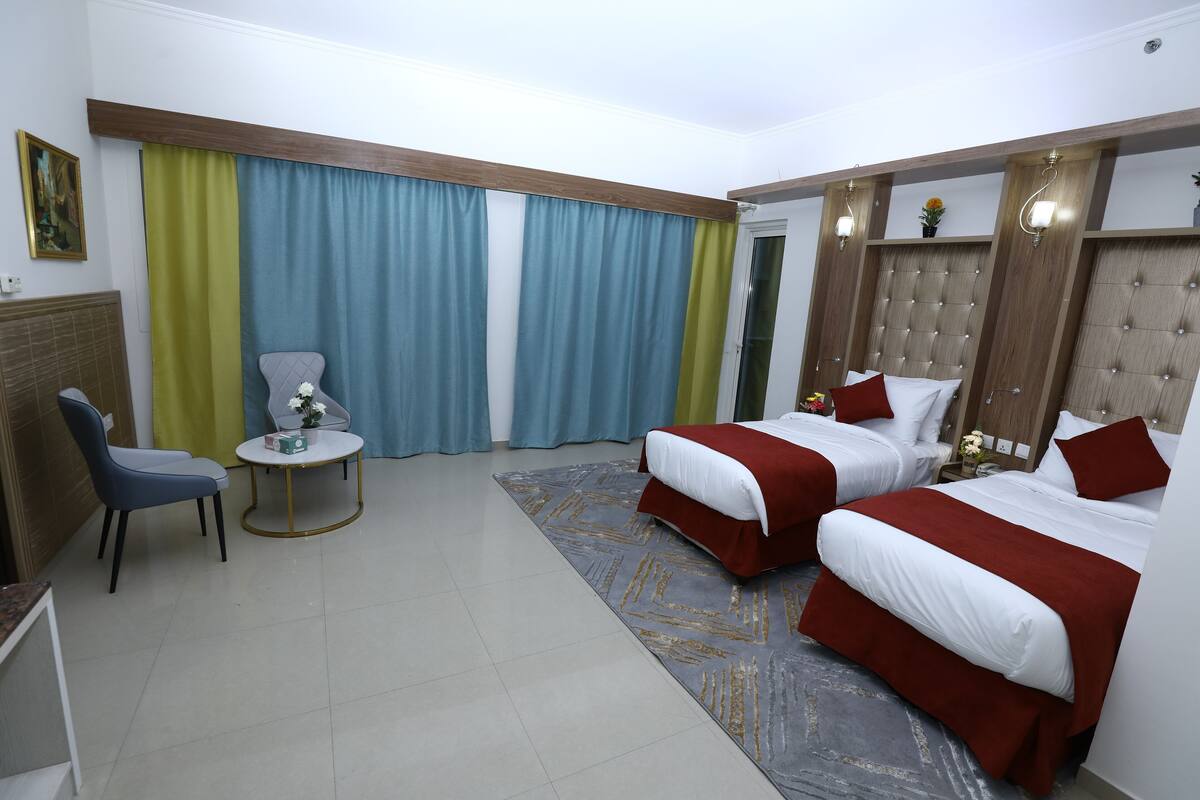 A hotel room with two beds, a small round table, a chair, and a large window with curtains.