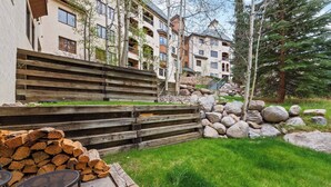 Un immeuble résidentiel avec un escalier en bois menant à un balcon, une pile de bois de chauffage et un espace herbeux avec une chaise en métal.
