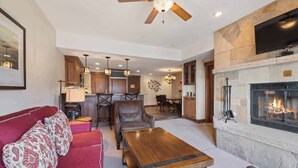 A living room with a fireplace, a ceiling fan, and a dining area with a kitchen in the background.