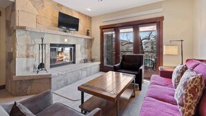 A living room with a stone fireplace, a flat-screen TV mounted above it, a ceiling fan, and a large window offering a view of the outdoors.