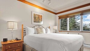 A bedroom with a large bed, wooden nightstand with a lamp, and a window with a view of trees. - The Charter at Beaver Creek C0405 (Beaver Creek)