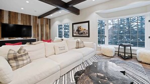 A modern living room with a large sofa, a coffee table, and a TV. There are windows with a view of trees and a fireplace. - The Charter at Beaver Creek E2405 (Beaver Creek)