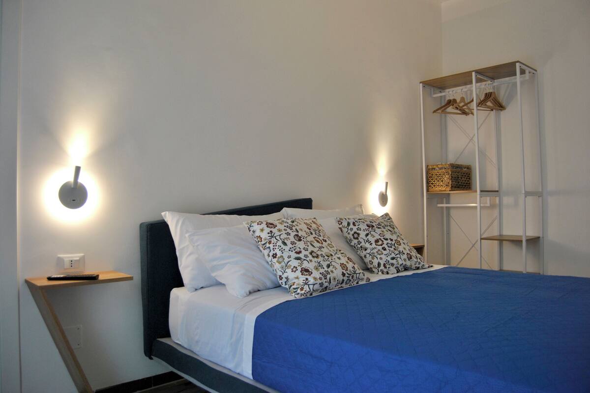 Cama bem arrumada com colcha azul e almofadas florais, mesa de cabeceira com controle remoto e prateleira na parede com cestos.