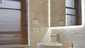 A bathroom with a wooden vanity, a white sink, and a mirror. There are towels stacked on a shelf below the vanity.