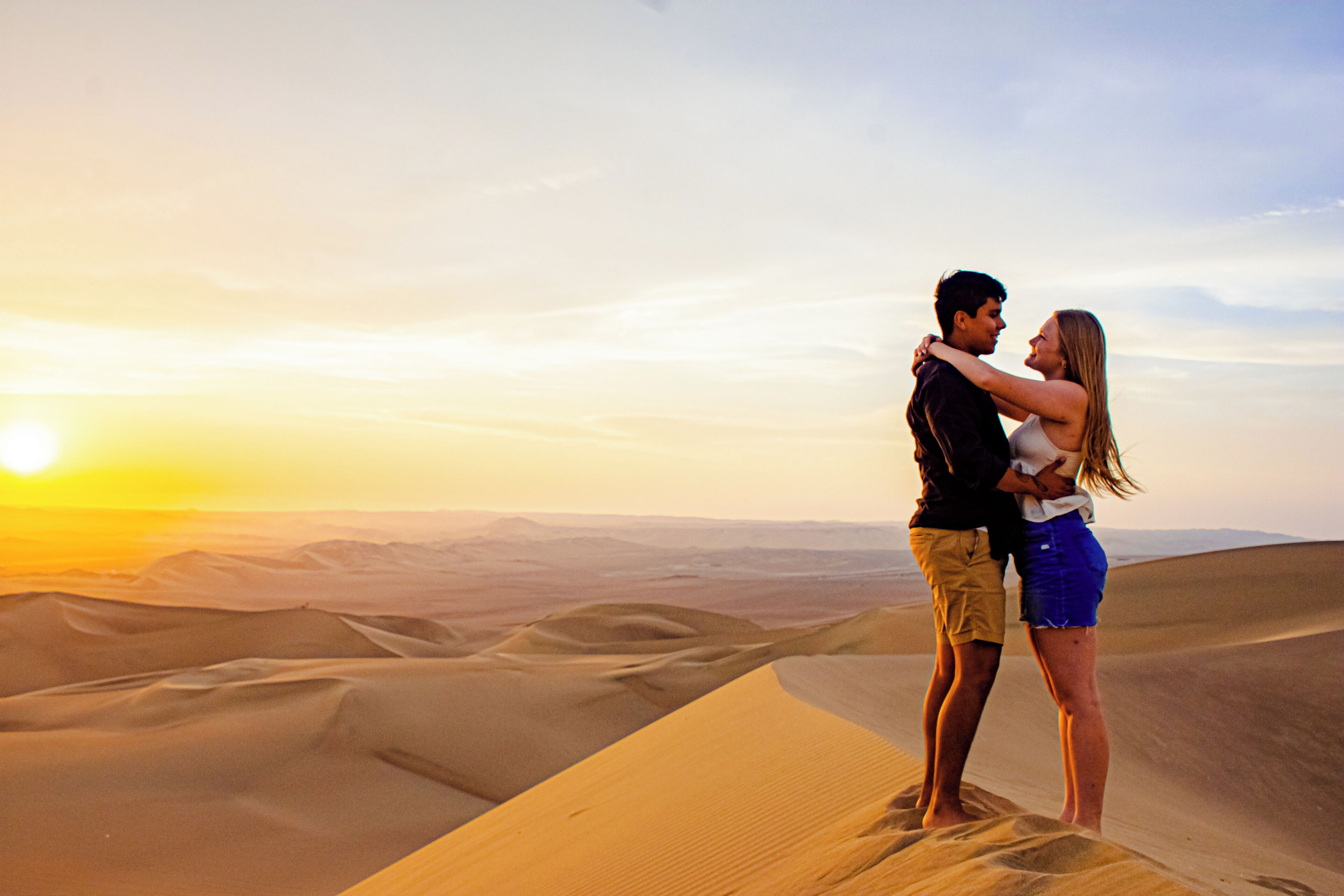 Una pareja se abraza en una duna de arena al atardecer.