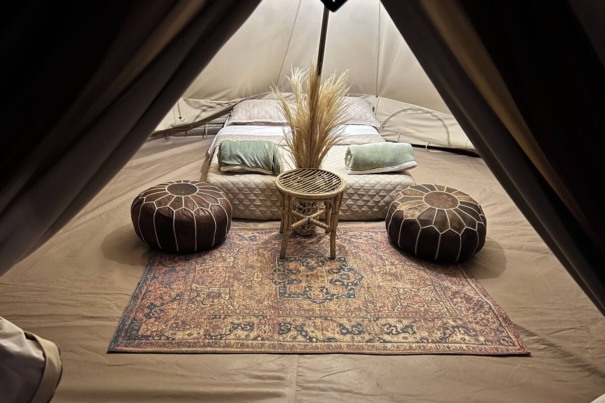 A tent with a bed, two pouf cushions, a woven chair, and a patterned rug.
