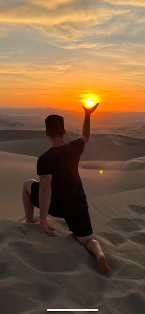 Una persona sentada en una duna de arena, con un atardecer de fondo.