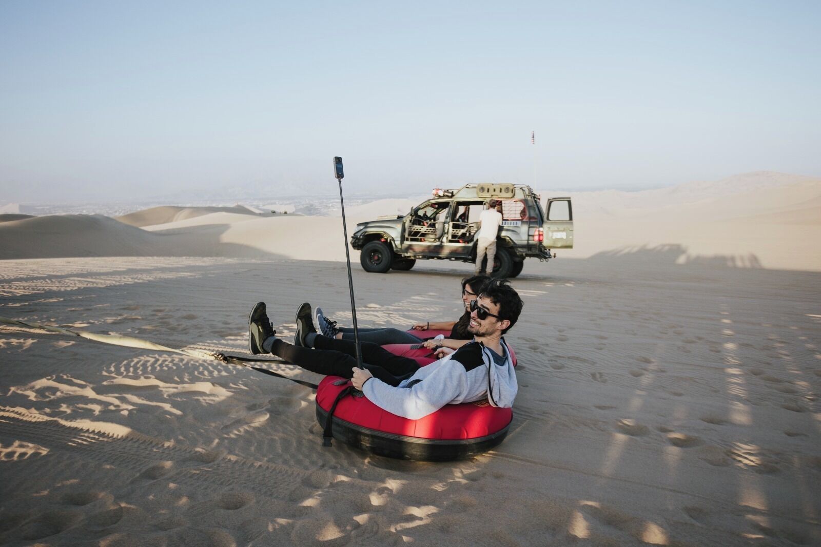 A person lying on a red inflatable tube in a desert with sand dunes in the background.