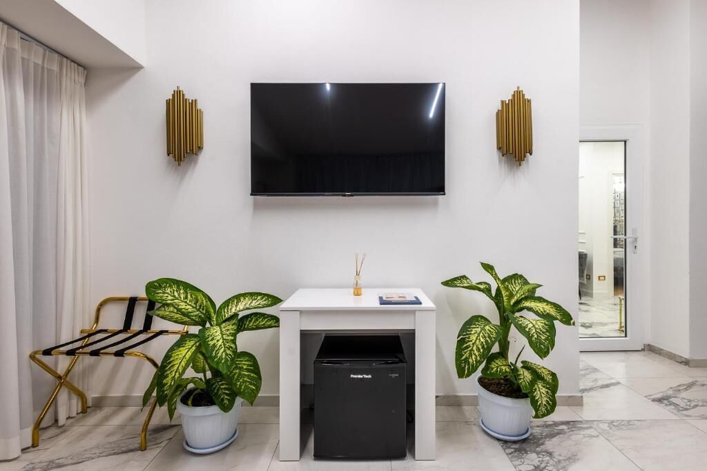 A modern living room with a flat-screen TV mounted on the wall, a small white table with a drawer, two potted plants, and a chair with a gold frame.