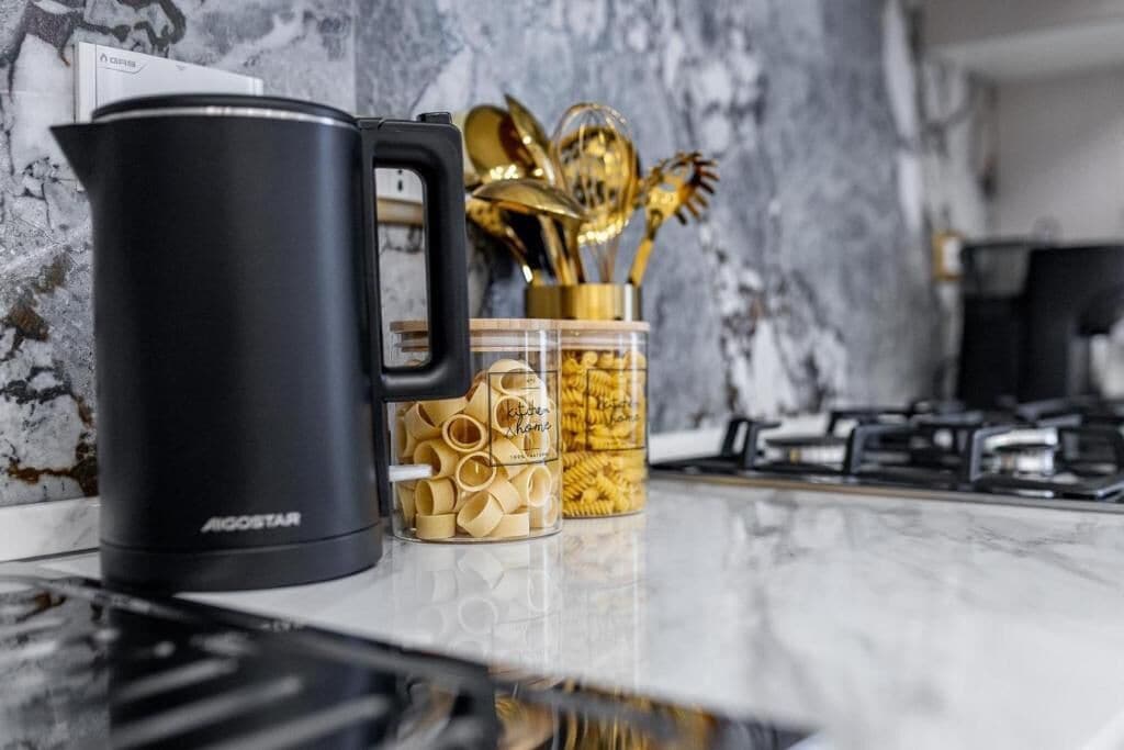 Eine Küchenarbeitsplatte mit einer schwarzen Kaffeemaschine, einem Glasbehälter mit Pasta und einem Set goldfarbener Küchenutensilien.