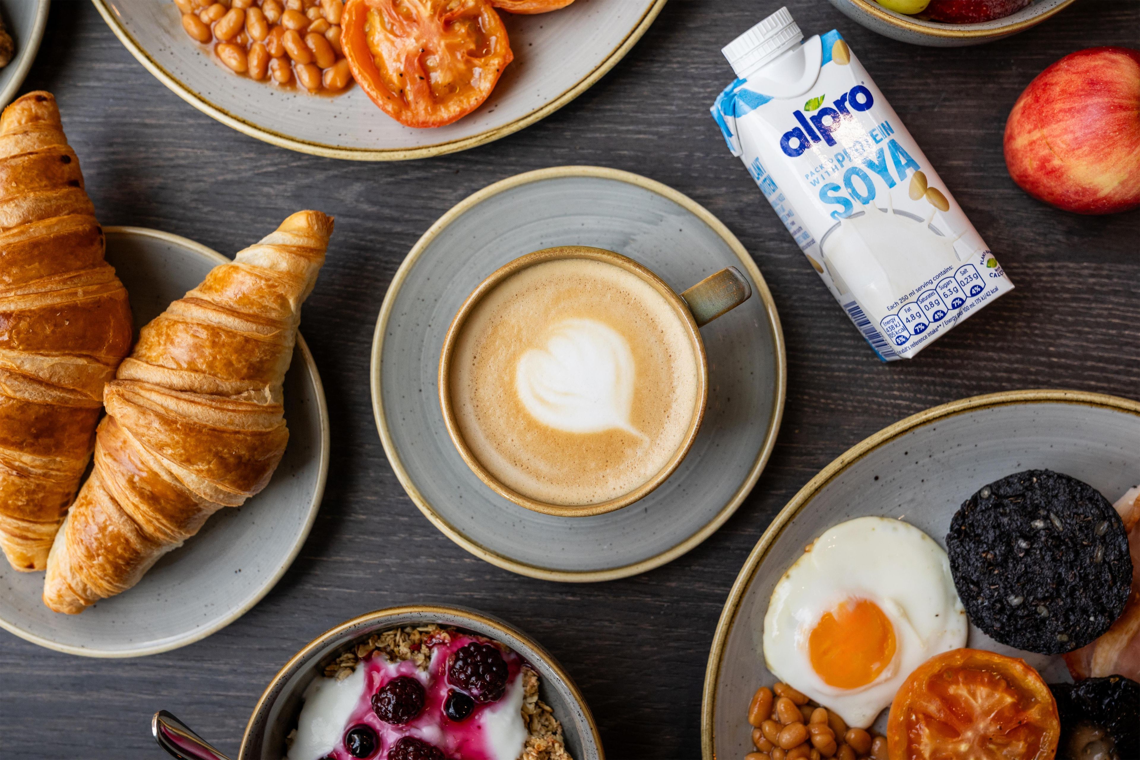 A breakfast setting with croissants, coffee, yogurt with berries, eggs, and a carton of almond milk.