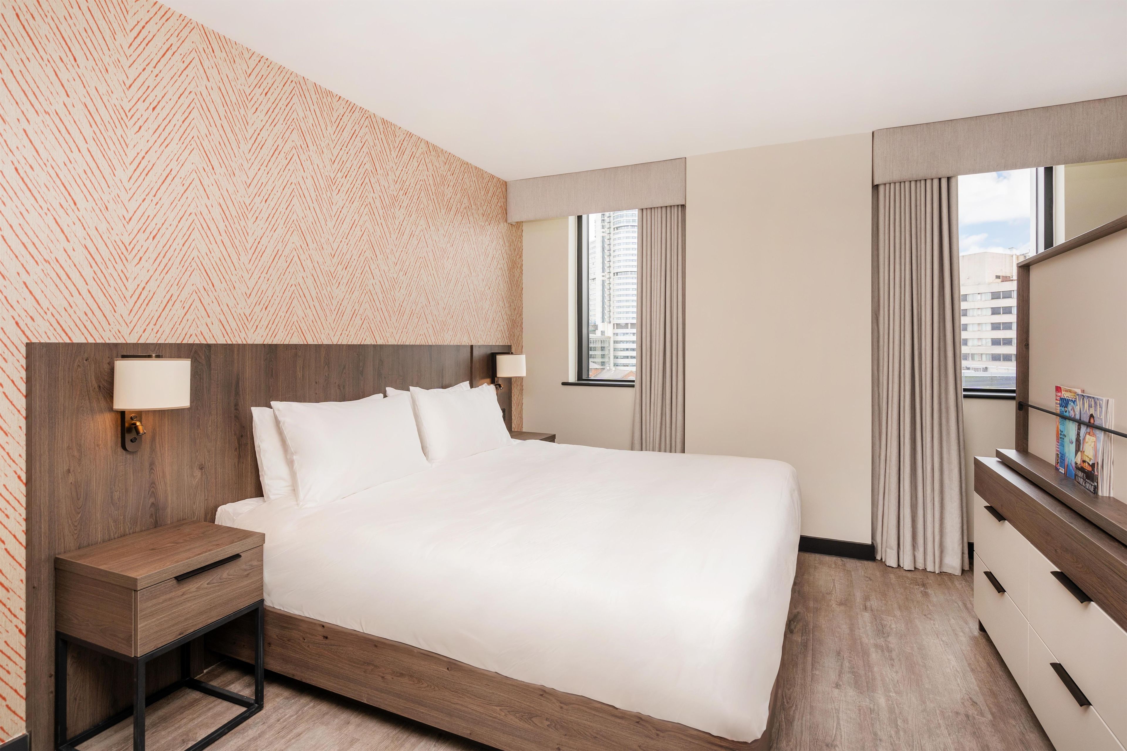 A hotel room with a large bed, bedside tables, a mirror, and a view of the city through the window.