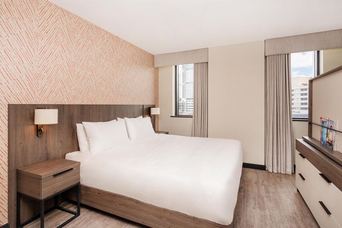 Hotellihuone, jossa on suuri sänky, yöpöydät, peili ja ikkunasta avautuva näkymä kaupunkiin.
