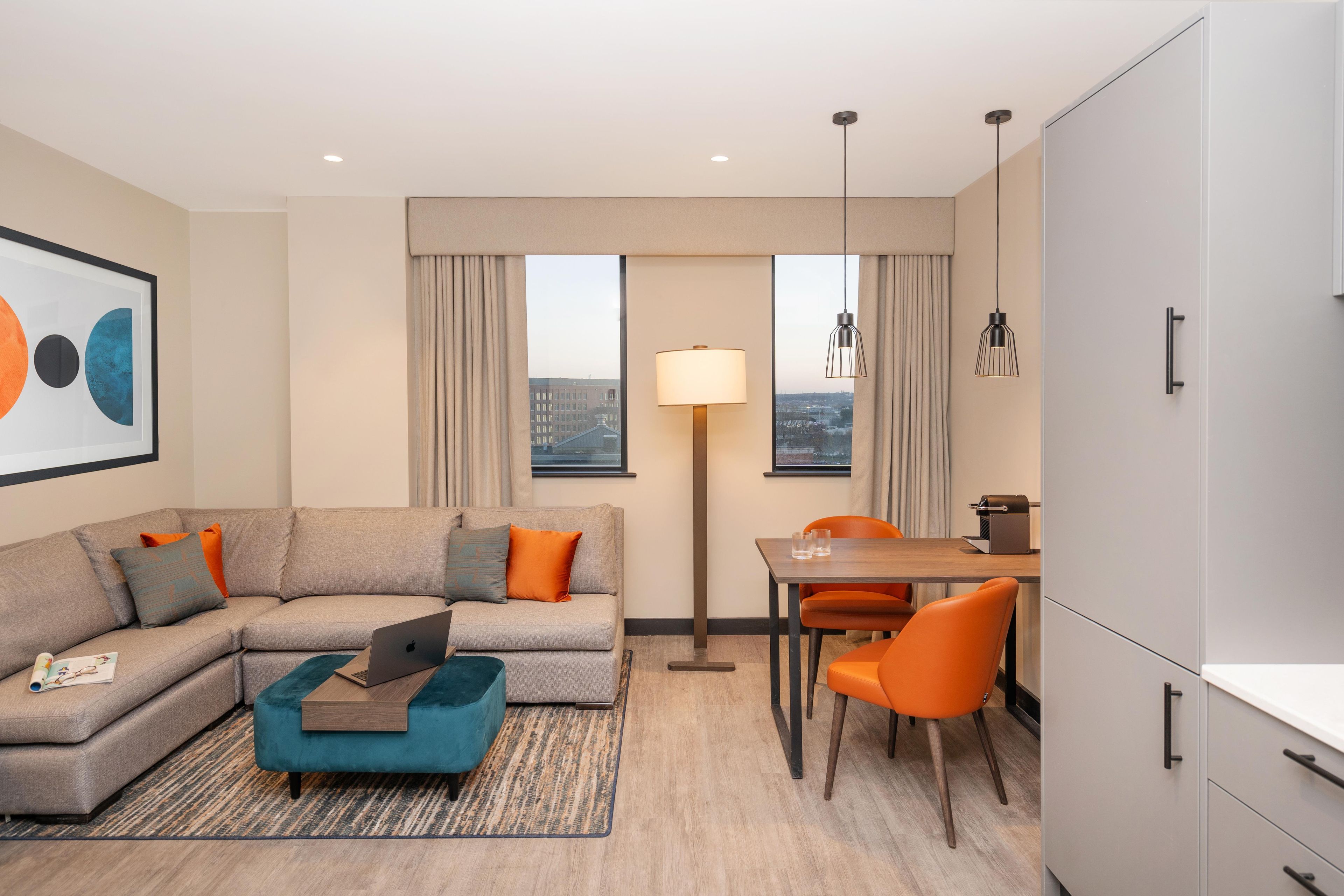 A modern hotel room with a sofa, a dining table with chairs, a lamp, and a view of the cityscape.