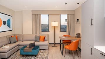 A modern hotel room with a sofa, a dining table with chairs, a lamp, and a view of the cityscape.