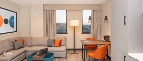 A modern hotel room with a sofa, a dining table with chairs, a lamp, and a view of the cityscape.