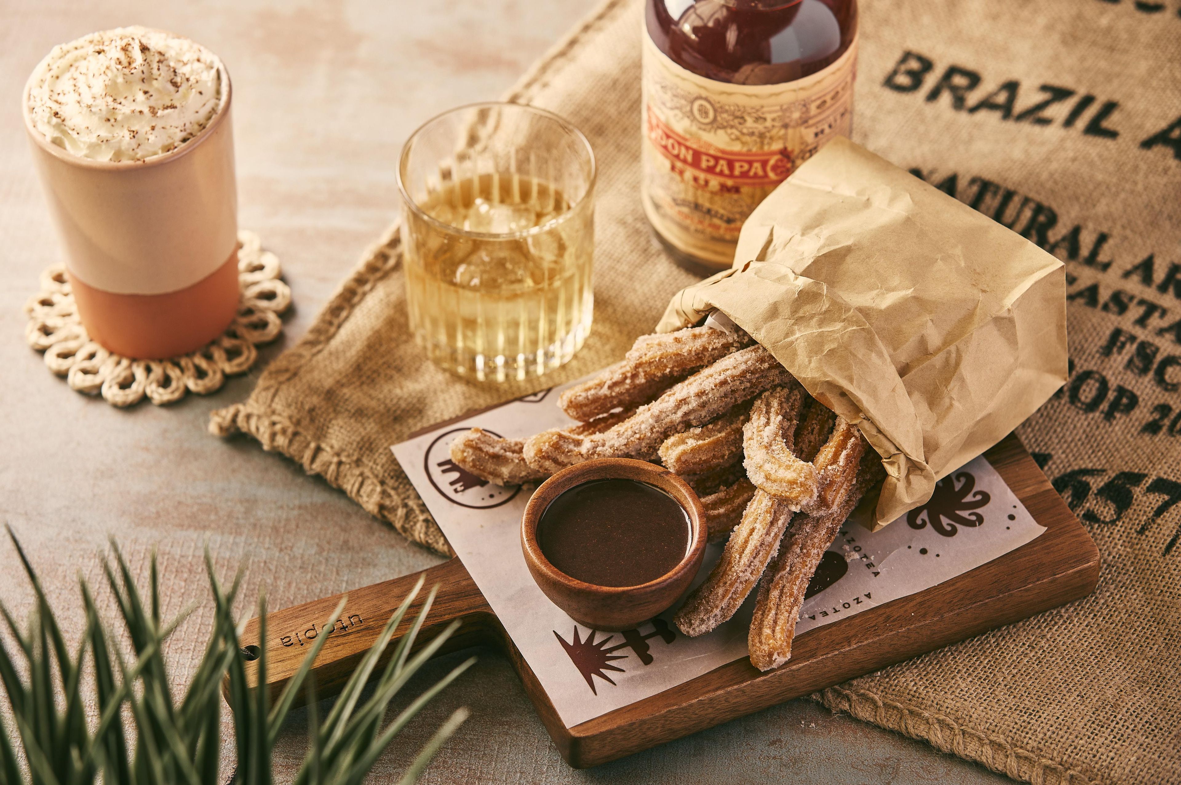 A wooden board with churros, a cup of hot chocolate, and a bottle of Coca-Cola.