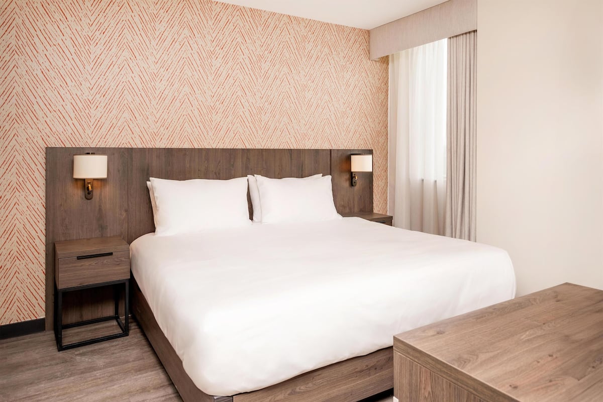 A hotel room with a large bed, two bedside tables with lamps, a wooden nightstand, and a wooden dresser.