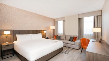 Habitación de hotel con una cama grande, un sofá, un escritorio con una computadora portátil y una silla. Hay dos lámparas de noche y un teléfono instalado en la pared.