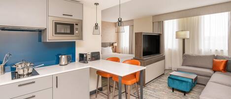 A modern kitchen with a microwave, kettle, and toaster. There is a dining area with a white table and orange chairs. A living area with a sofa and a TV is visible.