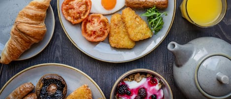 Un desayuno con platos de huevos, frijoles, tomates y tostadas, además de un croissant y un bol de fruta.