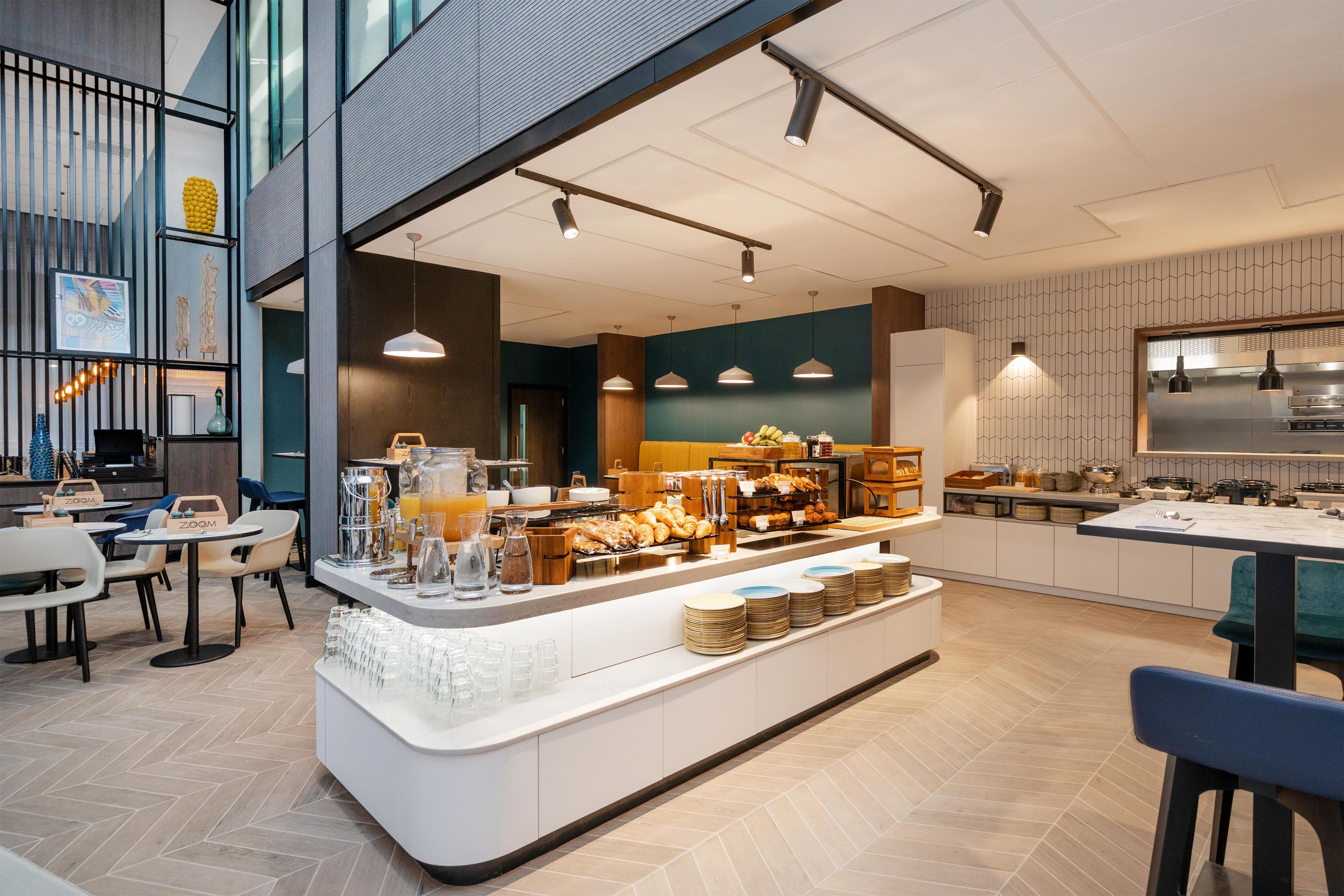 Un café modern amb un mostrador central que mostra aliments per esmorzar, zona de seients amb taules i cadires, i una cuina al fons.