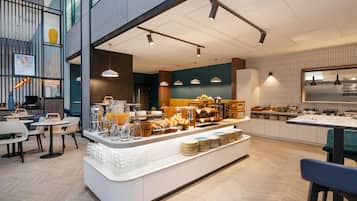 Un café modern amb un mostrador central que mostra aliments per esmorzar, zona de seients amb taules i cadires, i una cuina al fons.