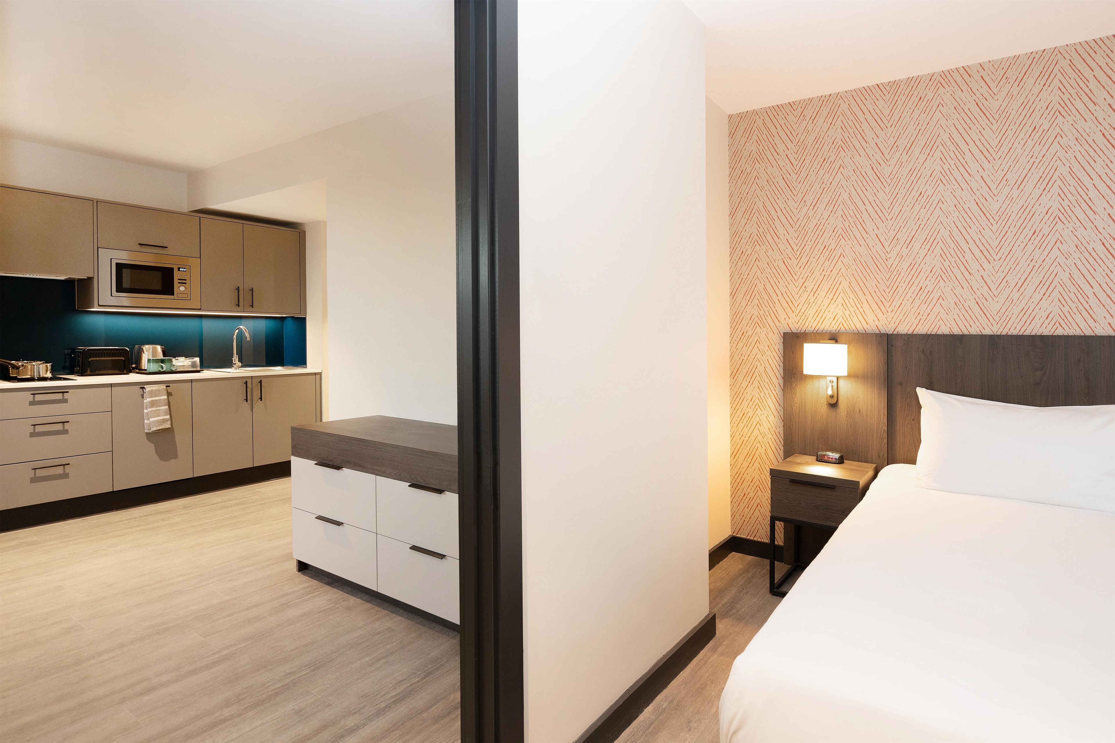 Een hotelkamer met een bed, nachtkastje met lamp, klein tafeltje met magnetron, en een kitchenette met kastjes en aanrecht.