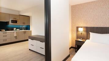 Een hotelkamer met een bed, nachtkastje met lamp, klein tafeltje met magnetron, en een kitchenette met kastjes en aanrecht.