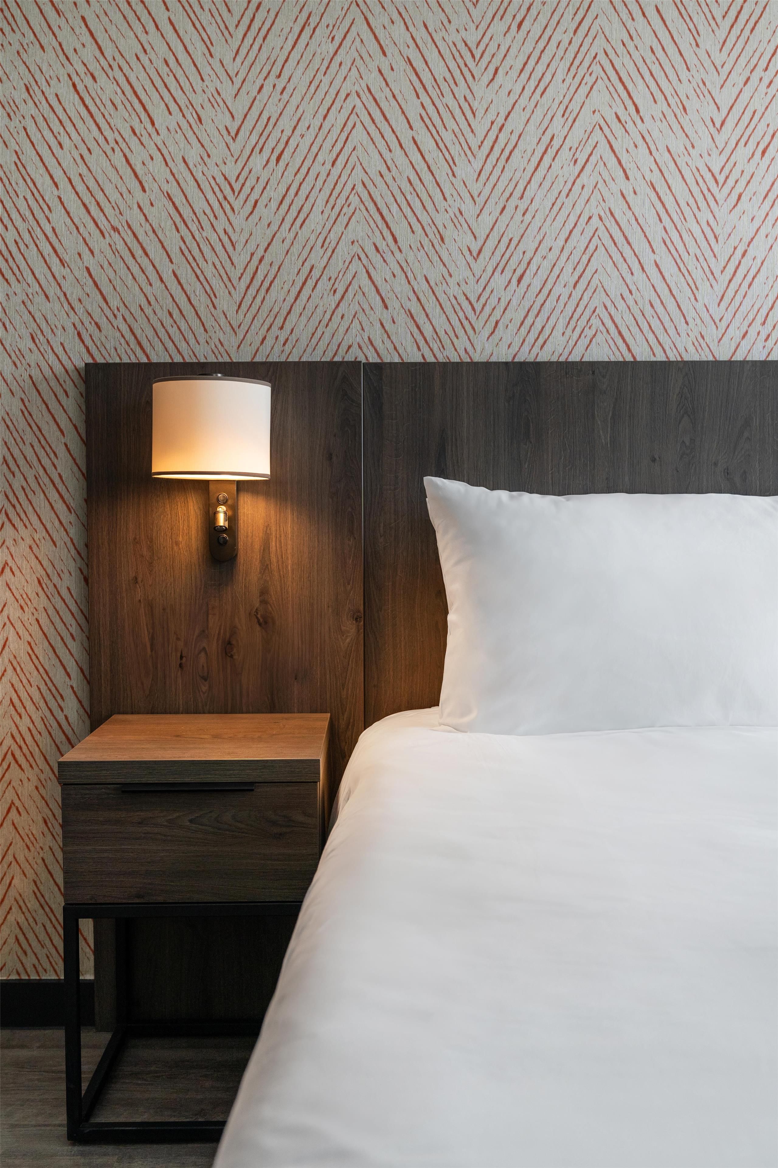 Habitación de hotel con cabecera de madera, mesita de noche con lámpara y una cama grande con ropa blanca de cama.