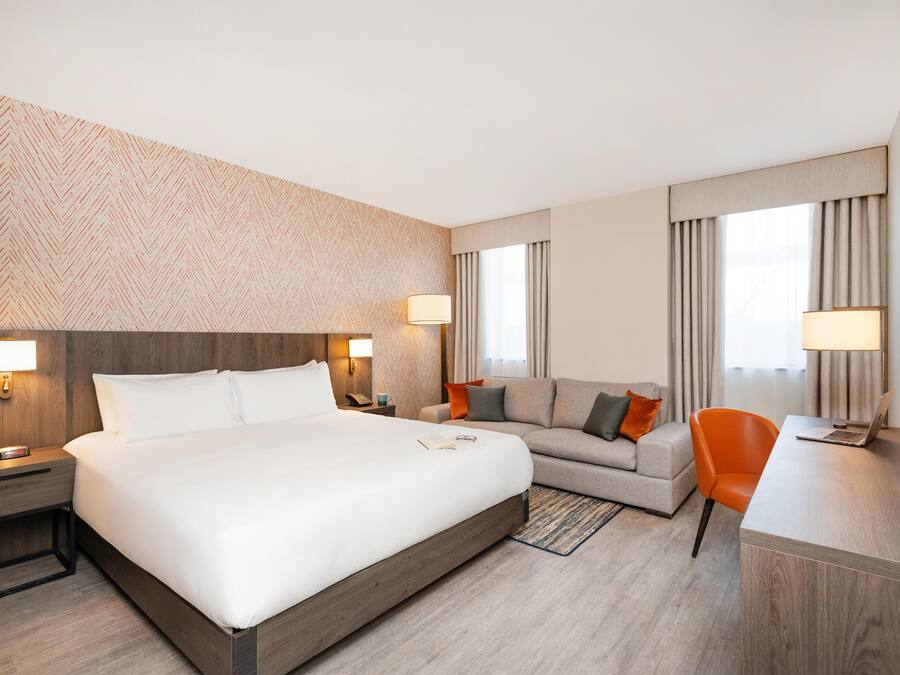 A hotel room with a large bed, a sofa, a desk with a laptop, and a chair. There are two bedside lamps and a wall-mounted phone.