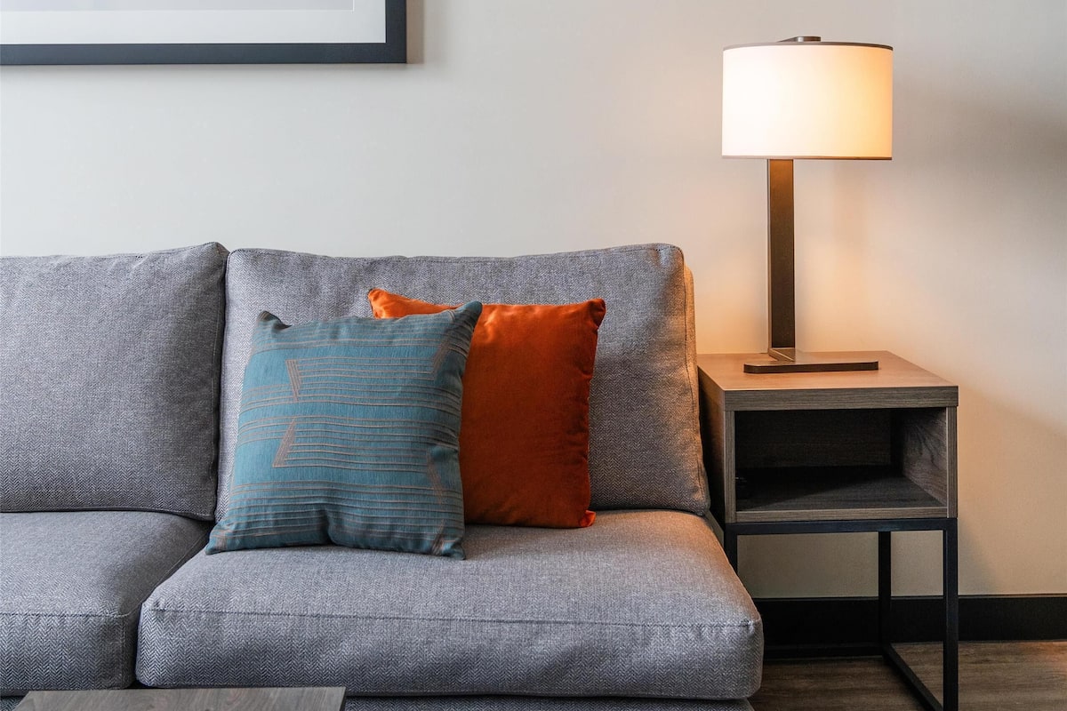 A modern living room with a grey sofa, a blue ottoman, a framed abstract painting, and a floor lamp.