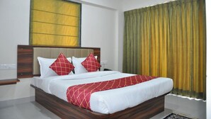 A hotel room with a wooden bed, white bedding, red and white pillows, a yellow window, and a green curtain. - Samrat Inn (Pune)