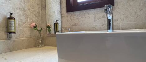 Une salle de bain avec une grande baignoire, un miroir et une fenêtre. On y trouve des articles de toilette sur le comptoir et un vase avec des fleurs.