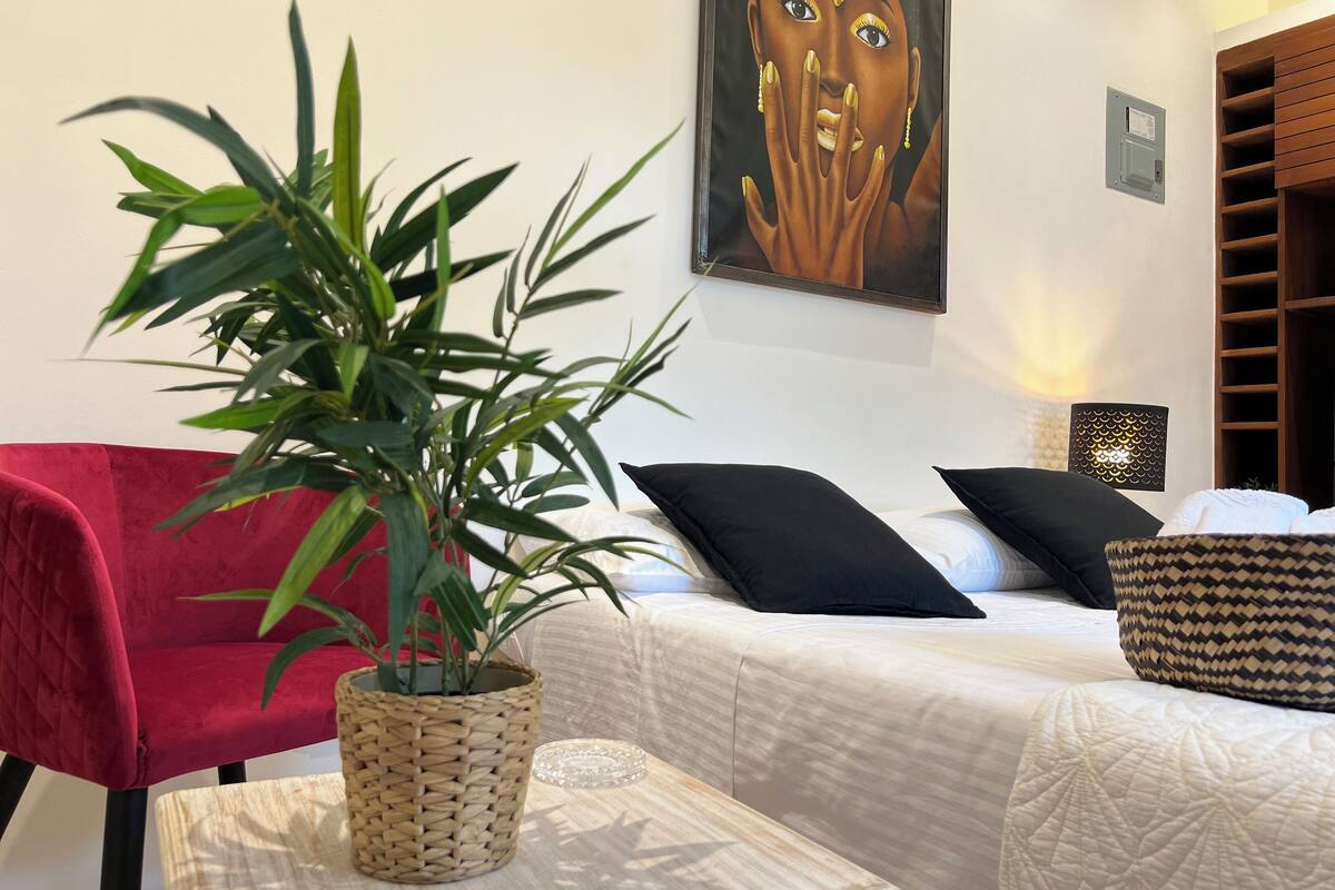 Una habitación con una silla roja, una cama con ropa de cama blanca, una planta en maceta y un cuadro en la pared.
