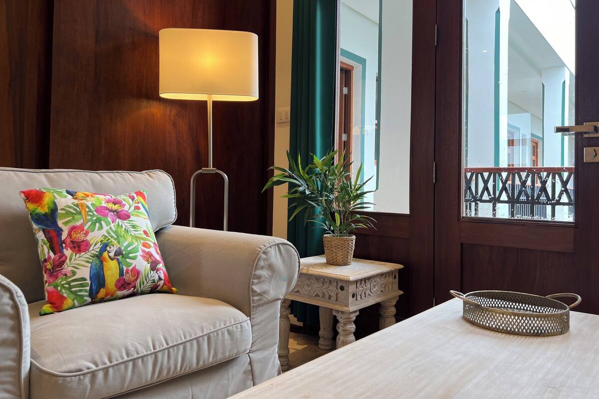 Un salón con sofá, lámpara, una mesita con una planta y una ventana con vista a un edificio.