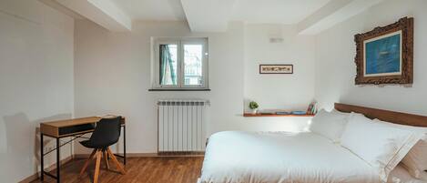 A bedroom with a wooden bed, a desk with a chair, a radiator, a window, and a framed picture on the wall.