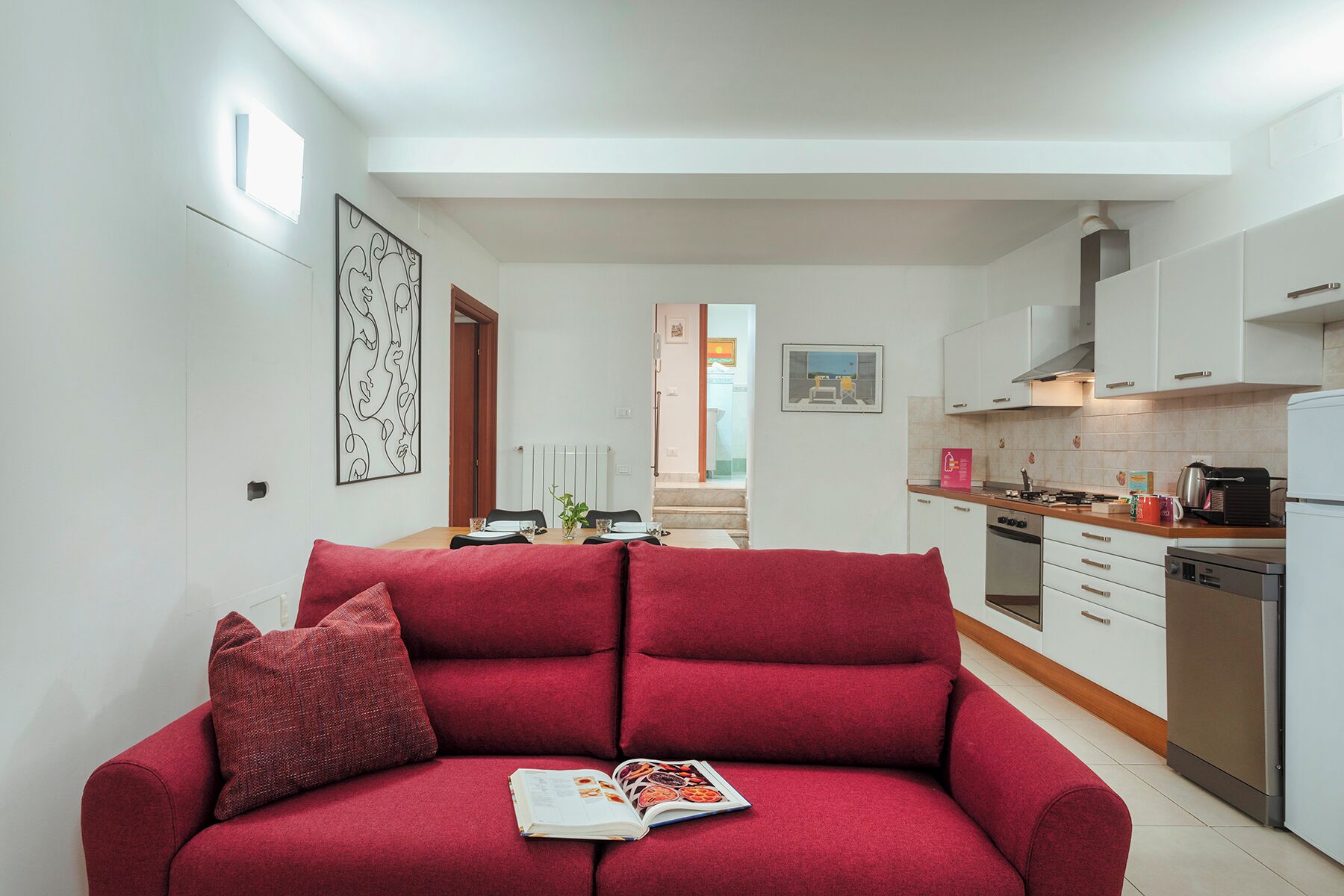 Una sala de estar moderna con un sofá rojo, un comedor y una cocina con gabinetes blancos y electrodomésticos de acero inoxidable.