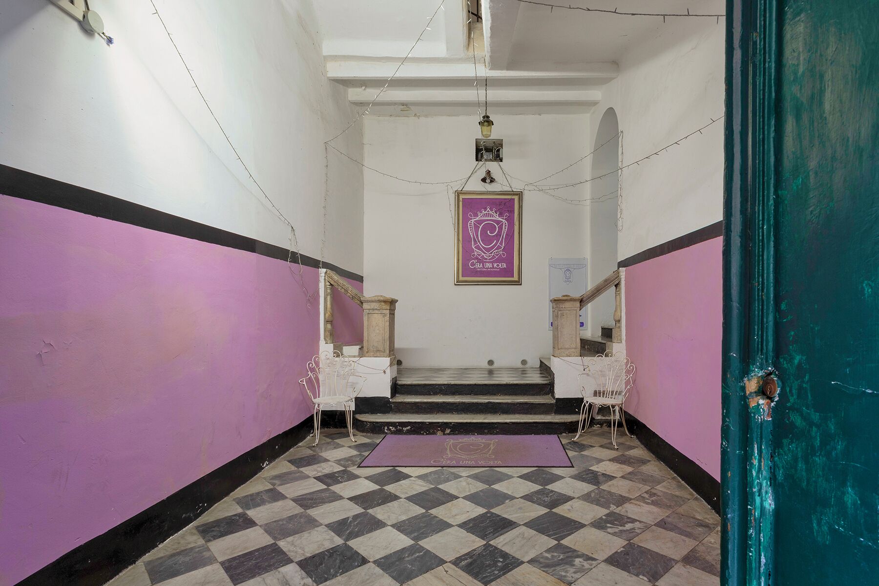 A narrow hallway with a checkered floor, white walls, and a purple accent wall. There are two white chairs and a purple mat at the bottom of a set of stairs.