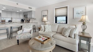 Ein modernes Wohnzimmer mit einer Couch, einem Hocker und Sesseln, eine Küche im Hintergrund und Blick auf die Stadt durch das Fenster.