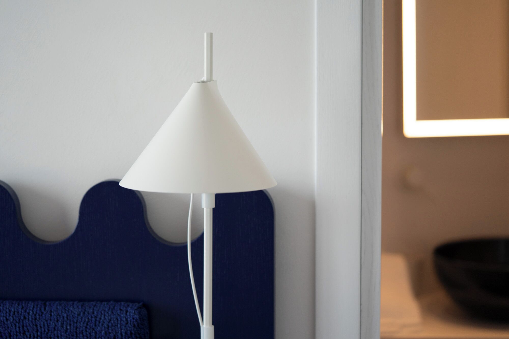 Une lampe de sol blanche, dotée d’un abat-jour conique, est placée contre un mur surmonté d’une tête de lit bleue recouverte de tissu.