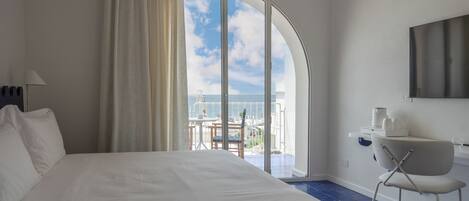 A hotel room with a large bed, a desk with a chair, a TV, and a balcony with a view of the sea.