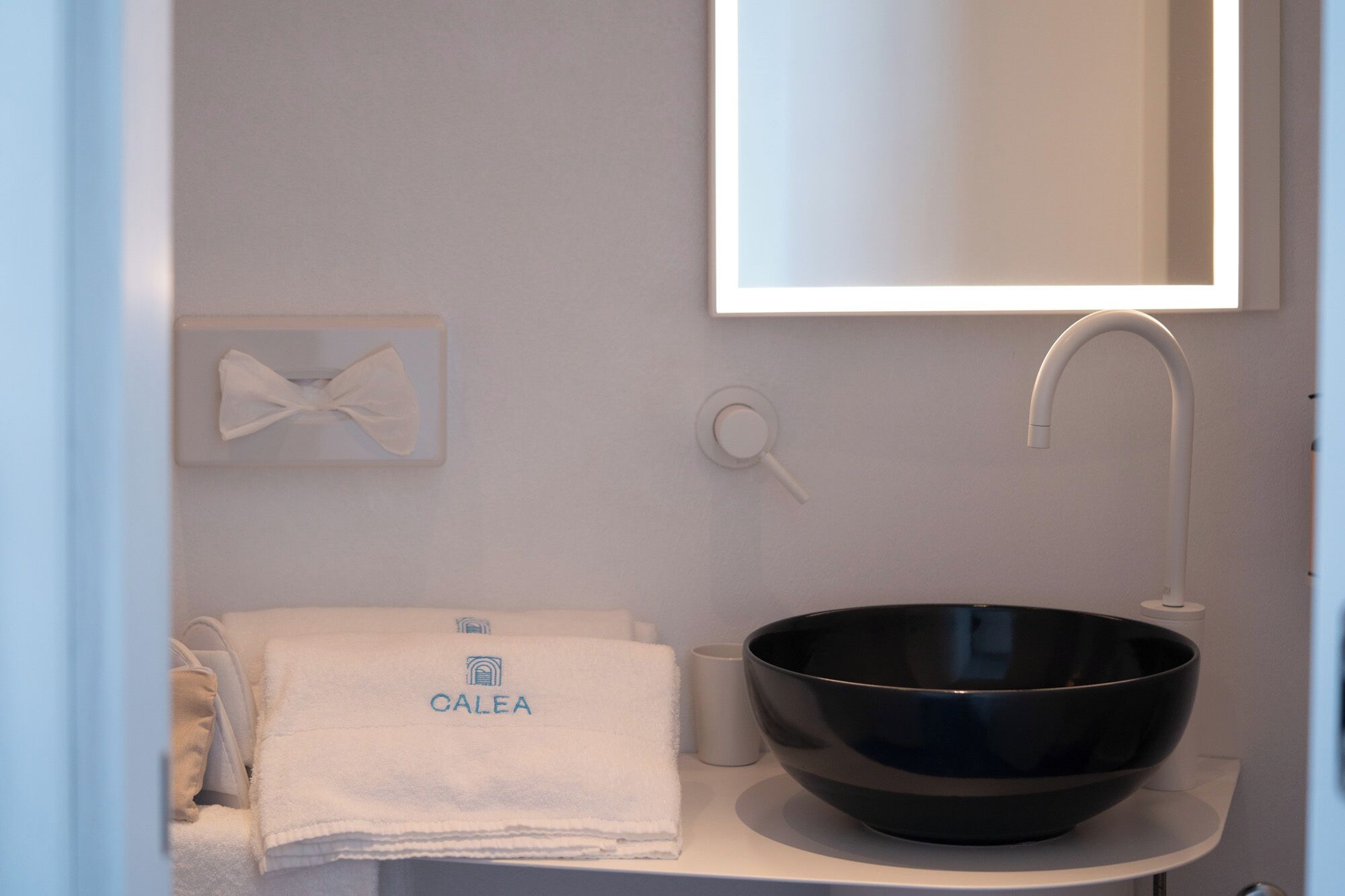 Une salle de bain avec un lavabo noir, une serviette ornée du logo « CALEA », une serviette blanche, une tasse blanche et un miroir.