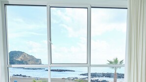 A large white bathtub with a glass door, positioned in front of a large window overlooking a beach with palm trees and rocky outcrops. - CALEA Boutique Hotel (Bacoli)